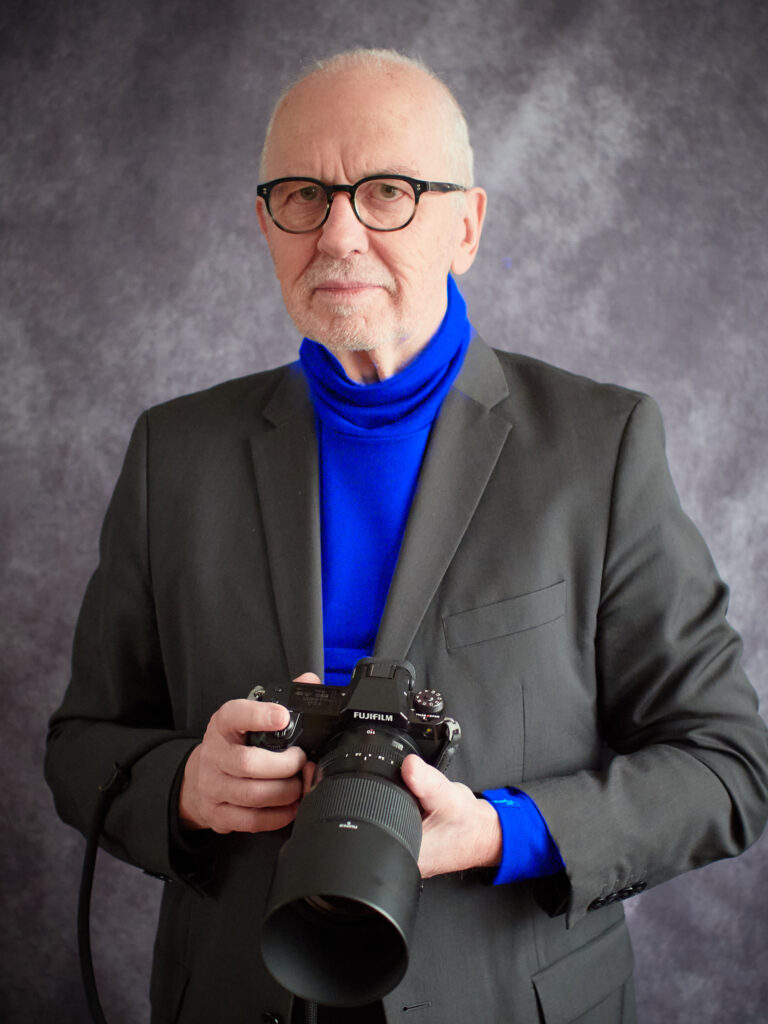 Peter Levin Schuster mit Kamera Der Fotograf Peter Levin Schuster hat einen schwarzen Anzug an und trägt eine Kamera um den Hals