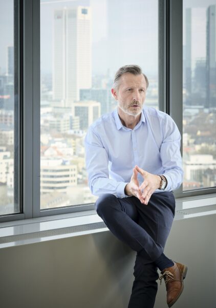 Mike sitzt am Fenster vor der Frankfurter Skyline, hält die Hände an den Fingern zusammen