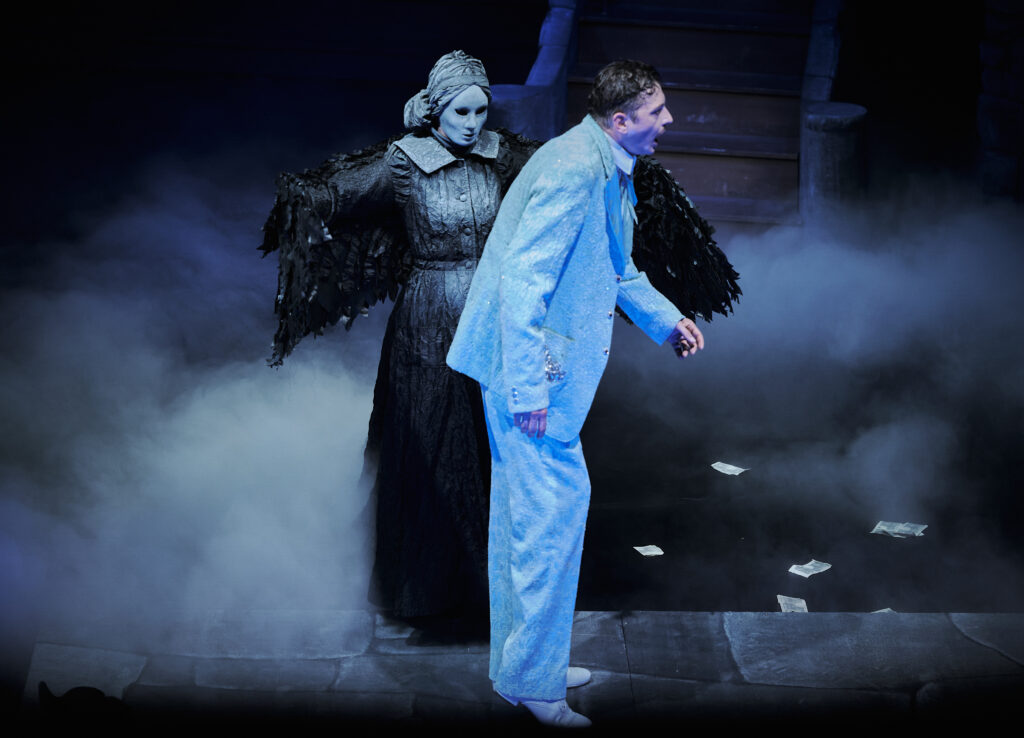 Dorian Gray im weißen Anzug und maskierte Todesfigur mit schwarzen Flügeln im Bühnennebel - Das Bildnis des Dorian Gray, Die Theater Chemnitz, Spielzeit 2025/26. Foto: Peter Levin Schuster