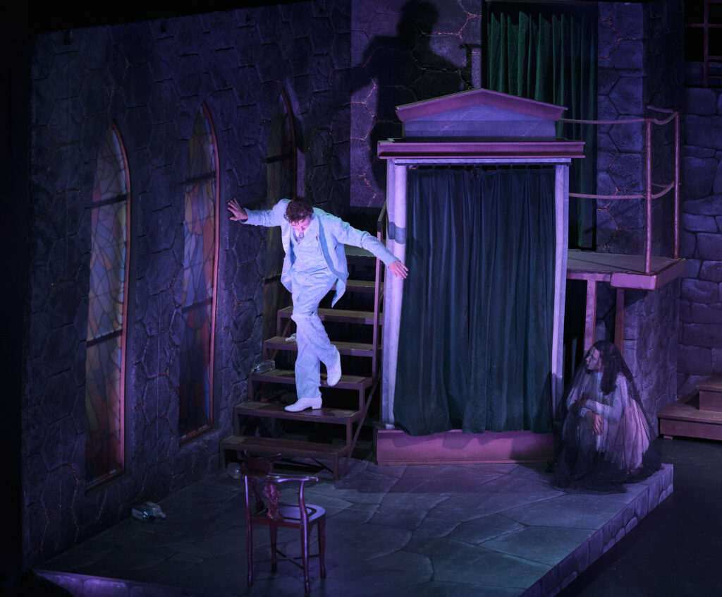Dorian Gray steigt die Bühnentreppe herab, kauernde Figur rechts im lila Bühnenlicht - Das Bildnis des Dorian Gray, Die Theater Chemnitz, Spielzeit 2025/26. Foto: Peter Levin Schuster